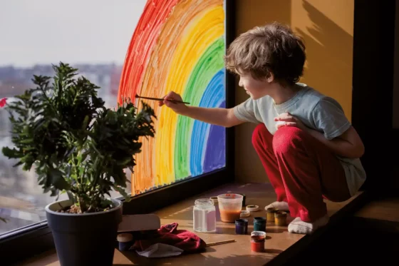 Junge sitzt auf der Fensterbank und malt einen Regenbogen ans Fenster